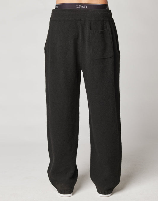 A person wears LIVE FAST DIE YOUNG’s Live Fast Boucle Pants in black, featuring a wide fit, high waist, Elastik-Bund with “LIV3ST.”, side pockets, and one front pocket. Made from 100% polyester, shown against a plain background.