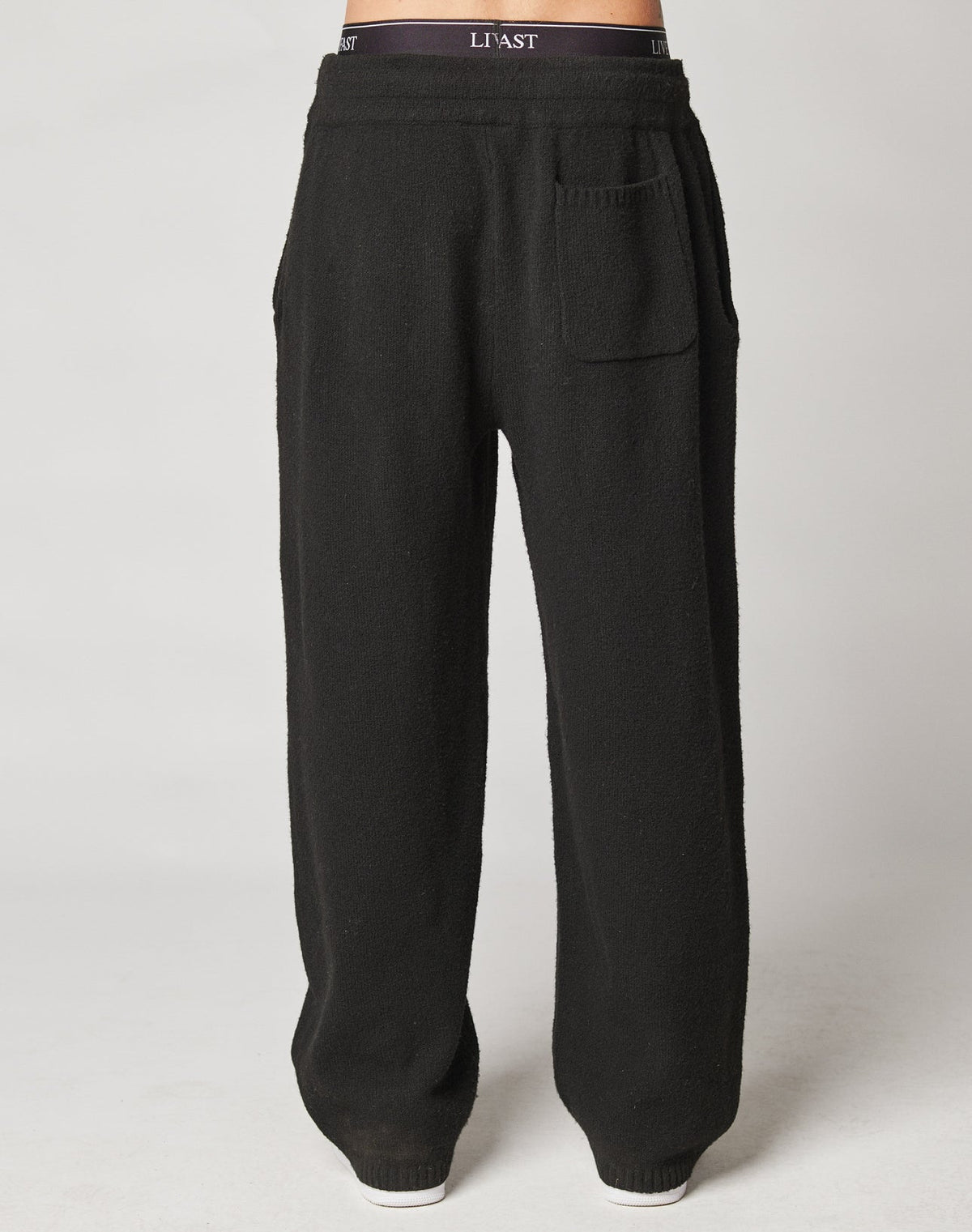 A person wears LIVE FAST DIE YOUNG’s Live Fast Boucle Pants in black, featuring a wide fit, high waist, Elastik-Bund with “LIV3ST.”, side pockets, and one front pocket. Made from 100% polyester, shown against a plain background.