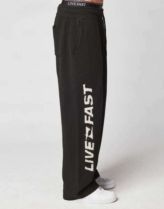 Someone wears black Live Fast Boucle Pants by LIVE FAST DIE YOUNG, featuring a wide fit, LIVE FAST in bold white letters down the side, an elastic waistband, and 100% polyester. Styled with white sneakers on a plain light background.