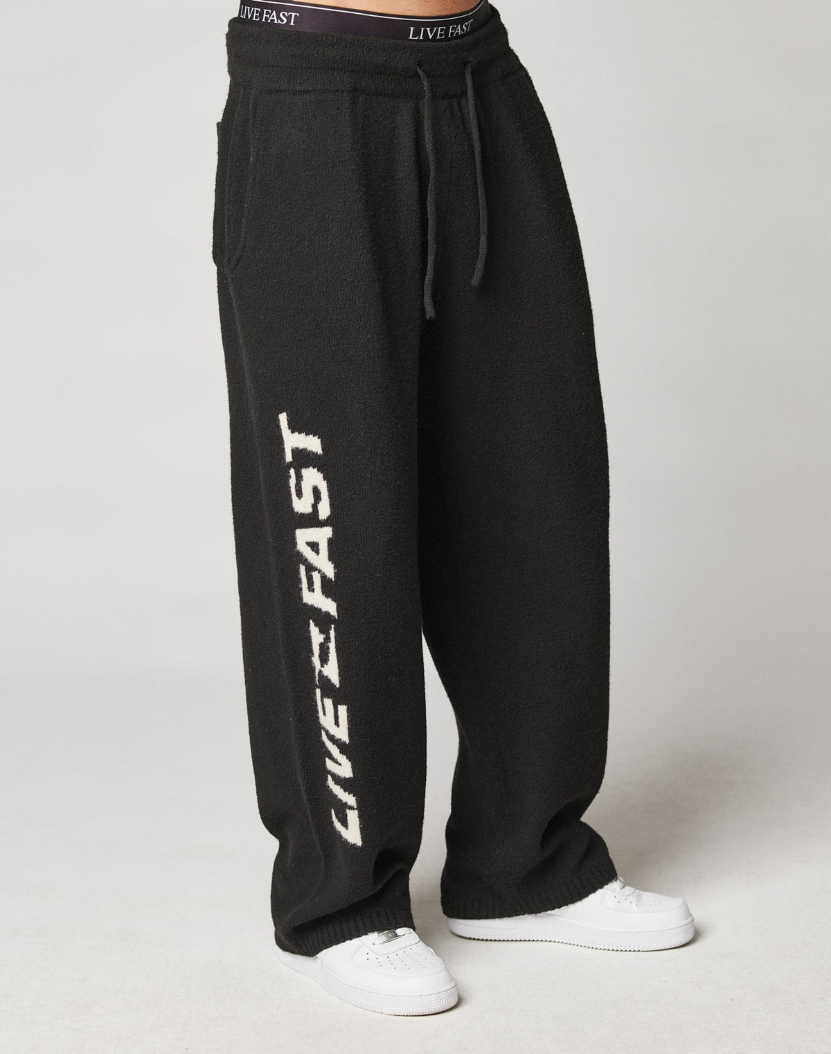 A person wears Live Fast Boucle Pants by LIVE FAST DIE YOUNG—loose black sweatpants with “LIVE FAST” in white on the left leg, wide fit, elastic drawstring waist, 100% polyester—styled with white sneakers against a light background.
