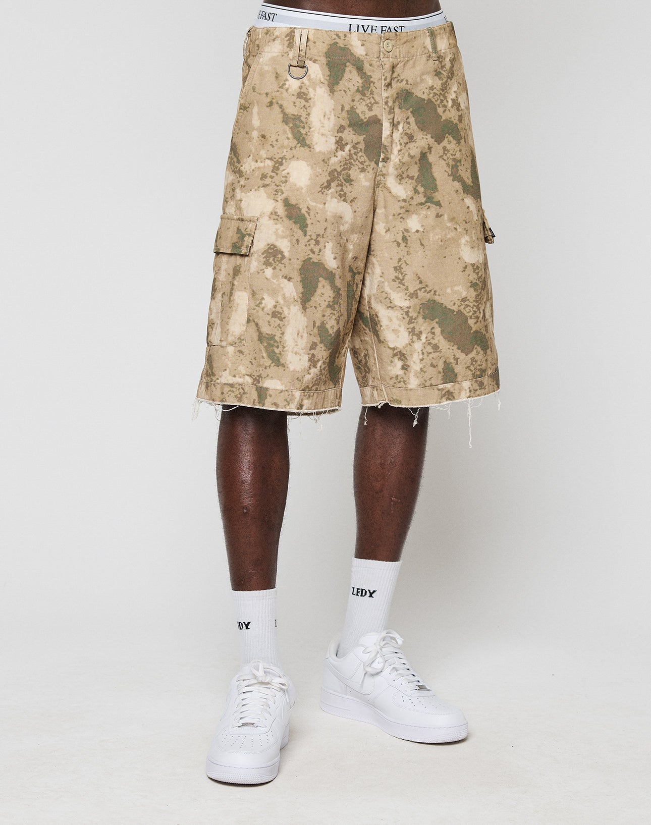 A person stands against a plain white background wearing LFDY Cargo Shorts by LIVE FAST DIE YOUNG in tan and green camo, made from 100% Baumwolle with frayed hems and a side pocket, plus white crew socks and sneakers. Model’s head not visible.