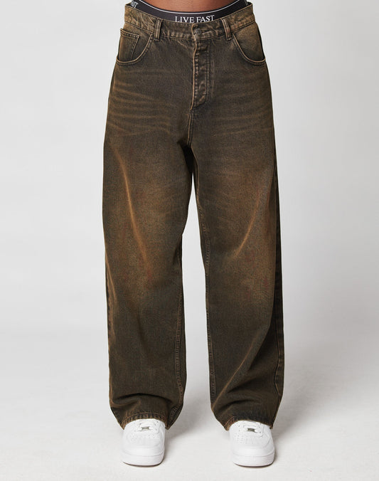 A person wears LFDY Baggy Denim by LIVE FAST DIE YOUNG, faded brown 100% Baumwolle jeans with gathered ankles, white sneakers, and a black waistband reading LIVE FAST. The background is plain and light-colored.