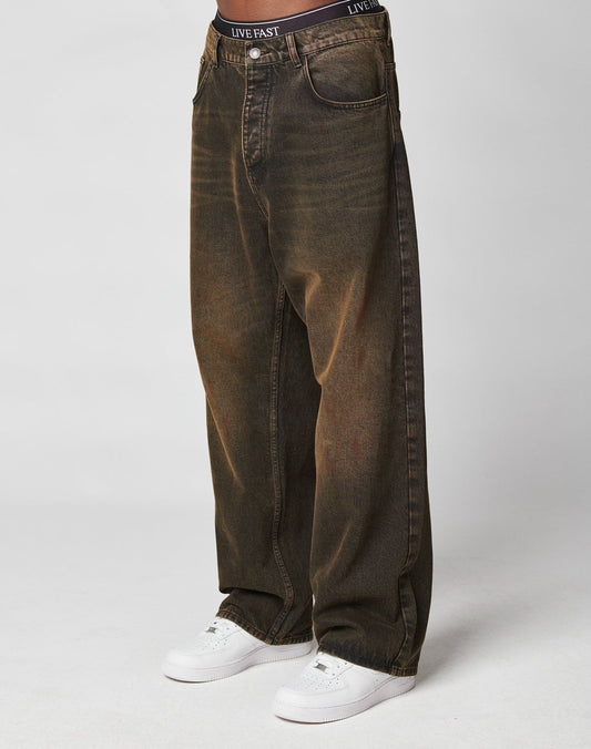A person wearing LIVE FAST DIE YOUNG’s LFDY Baggy Denim—loose, dark brown jeans with a faded wash and baggy fit—paired with white sneakers and black underwear reading LIVE FAST, standing against a plain white background.