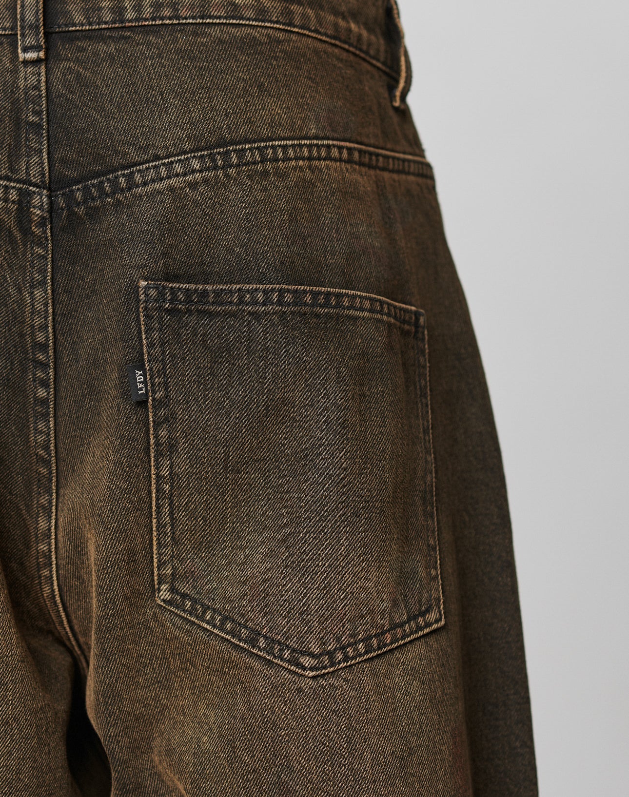 Close-up of the back pocket of LFDY Baggy Denim by LIVE FAST DIE YOUNG, featuring faded brown denim, baggy fit, visible stitching details, and a small black flag label on the pocket side.