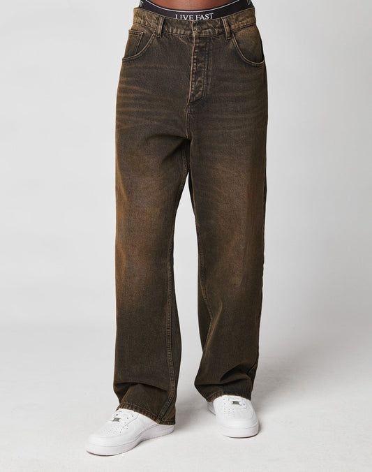 A person in loose-fitting, faded brown LFDY Baggy Denim by LIVE FAST DIE YOUNG, paired with white sneakers, stands against a plain light background. The waistband of their black LIVE FAST underwear is visible above the jeans.