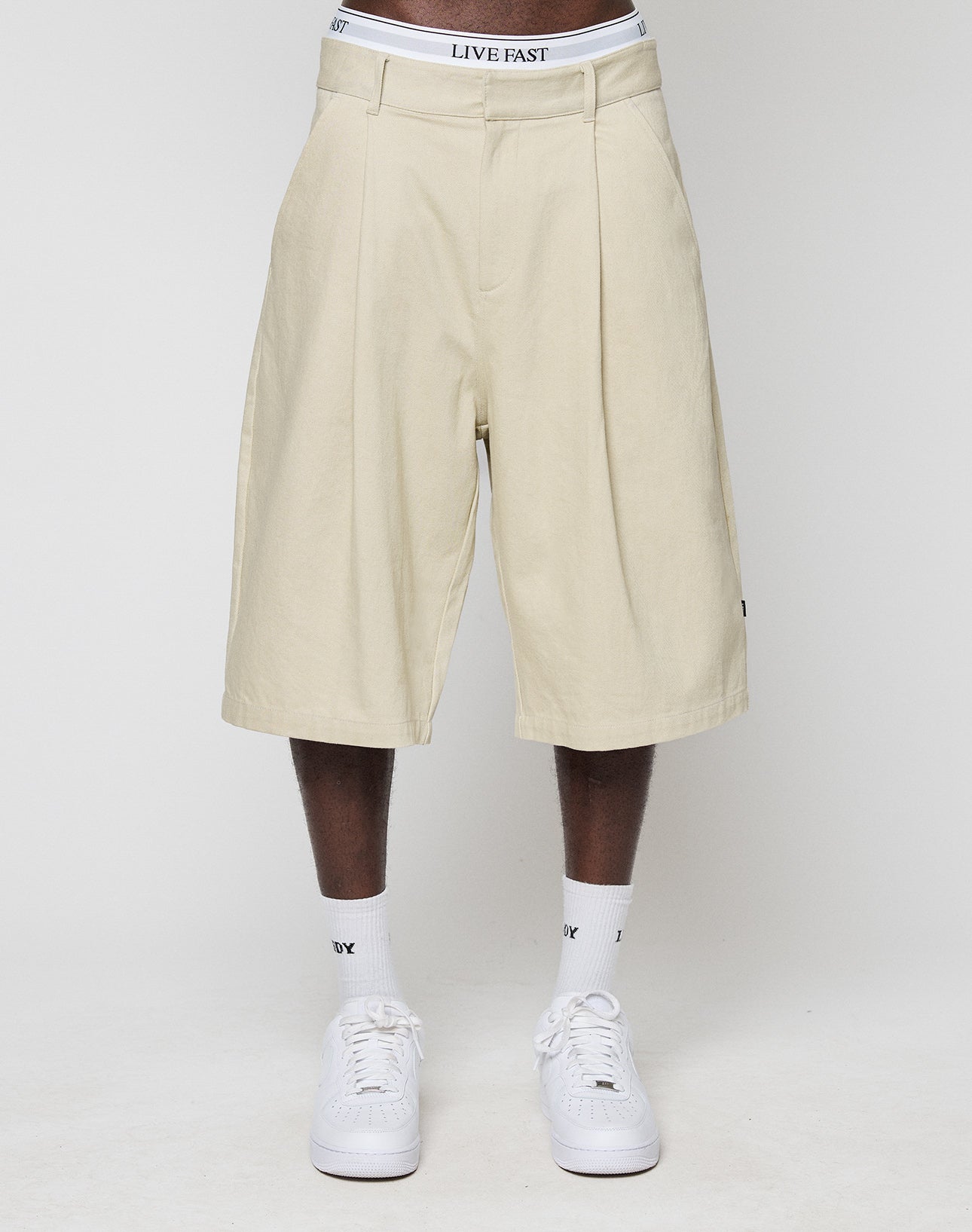 A person wears beige, knee-length LFDY Twill Shorts by LIVE FAST DIE YOUNG with an oversize fit and wide straight leg, paired with white sneakers, socks, and a visible waistband reading LIVE FAST, standing against a plain white background.