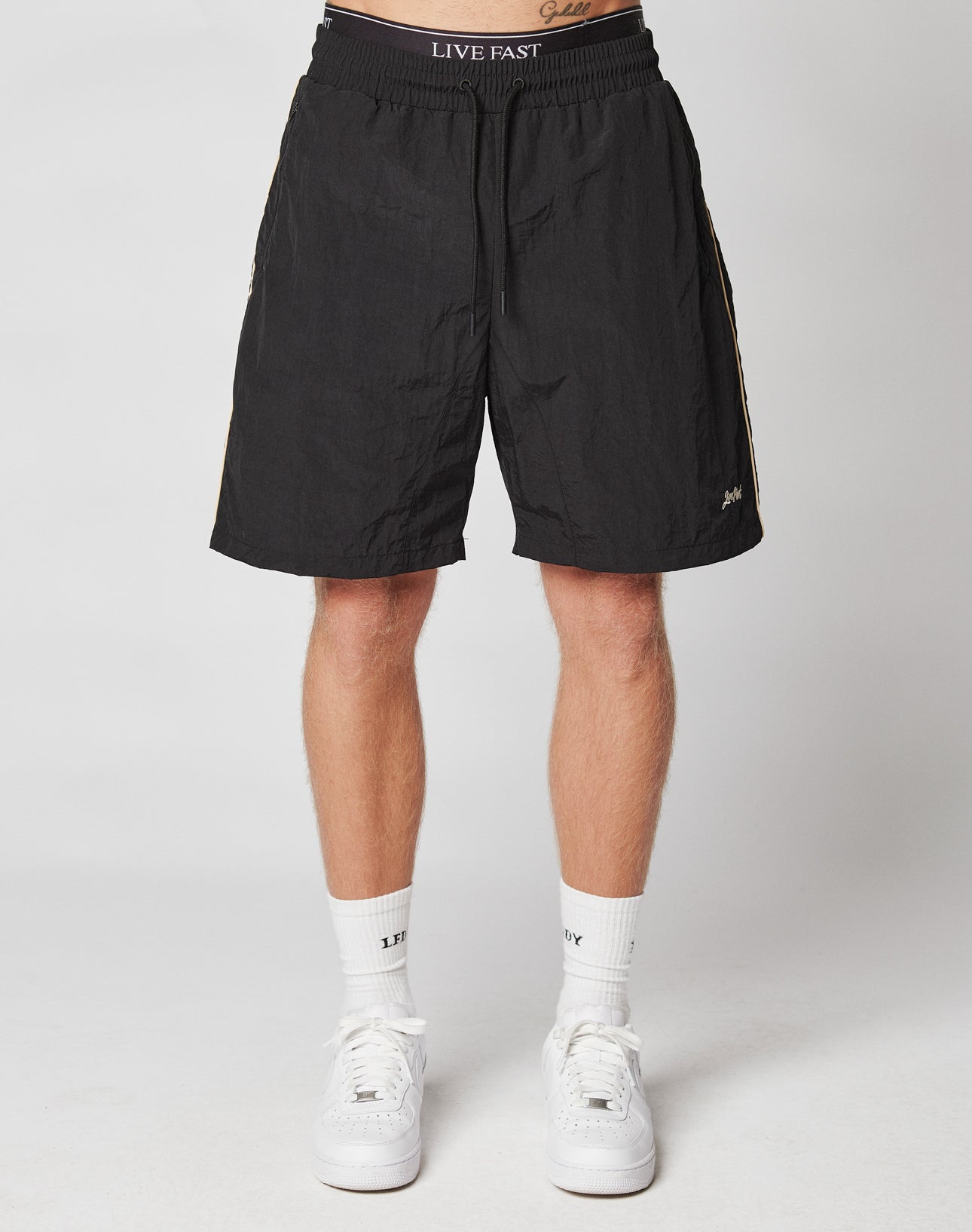 A person’s lower body is shown wearing black LIVE FAST DIE YOUNG Beach Shorts with an Elastik-Bund and mesh inlay, plus white socks and sneakers. The waistband reads “LIVE FAST.” They stand against a plain white background.