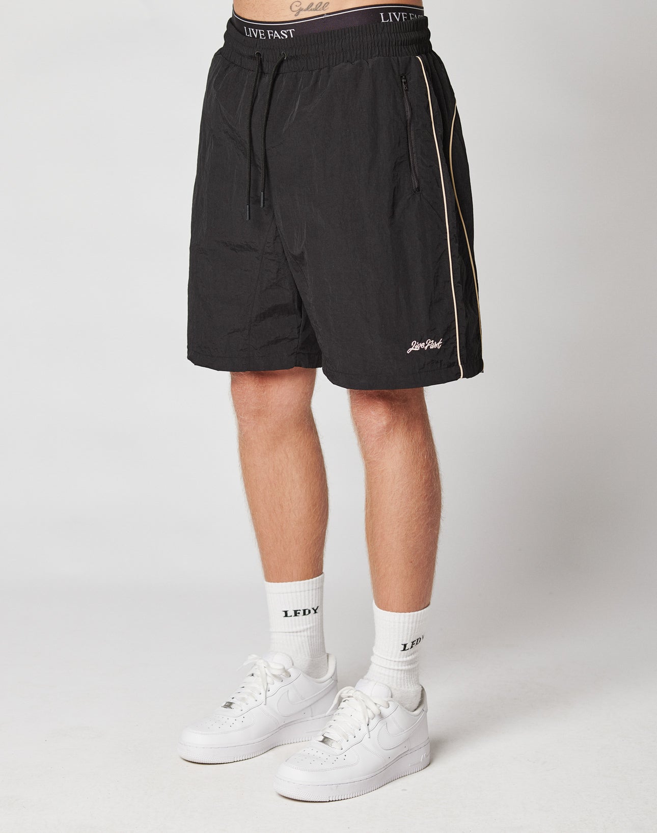A person wearing LIVE FAST DIE YOUNG Beach Shorts, white crew socks with LFDY text, and white Nike sneakers stands against a plain light background.