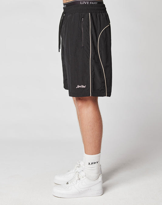 A person stands sideways wearing black LIVE FAST DIE YOUNG Beach Shorts with elastic waistband and white piping, white crew socks labeled LEDY, and white sneakers against a plain light background.