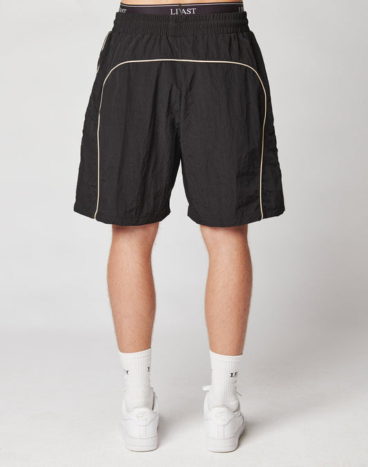 Someone is standing with their back to the camera against a plain light background, wearing LIVE FAST DIE YOUNG Beach Shorts in black with beige piping, paired with white socks and white sneakers.