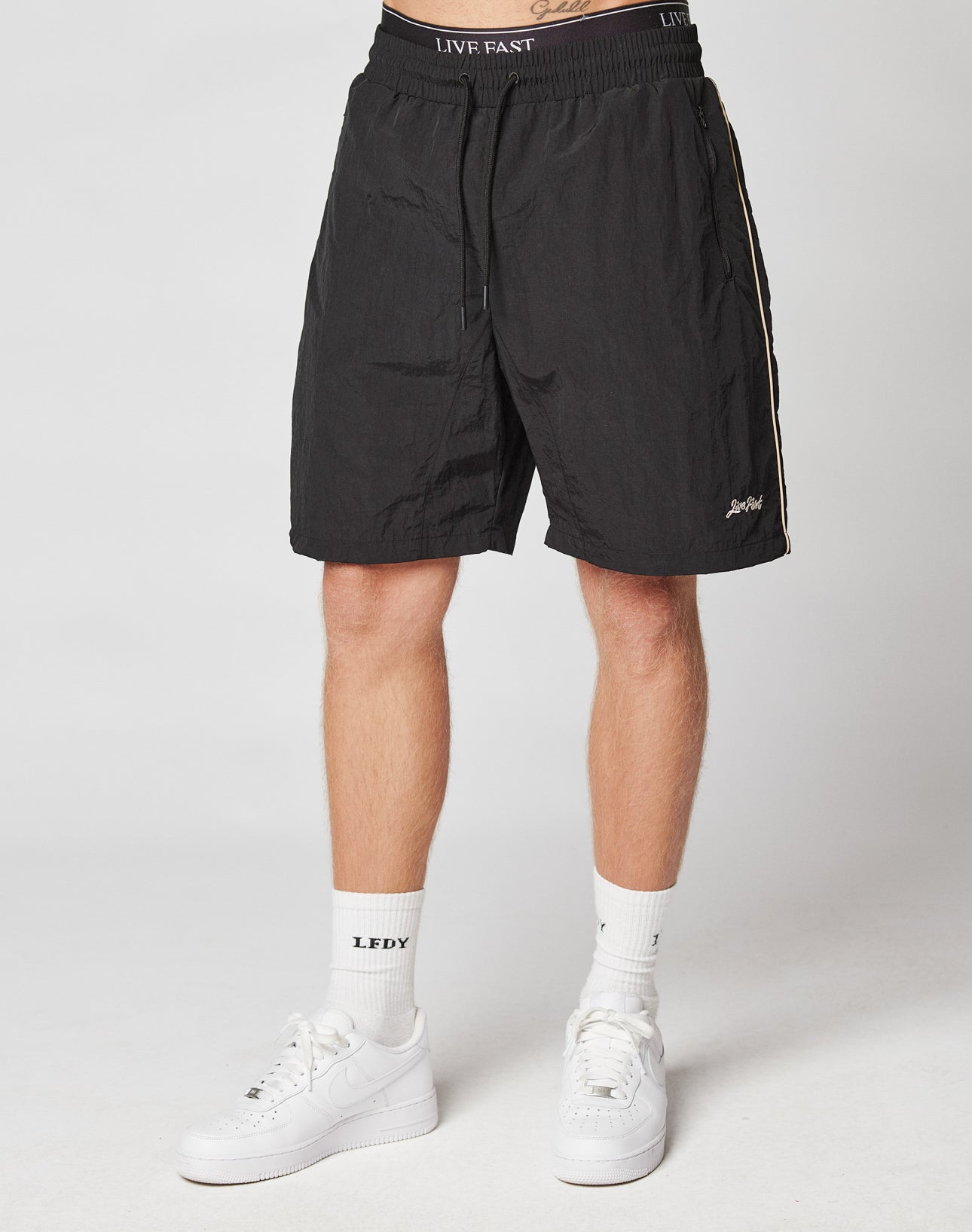 A person in black LIVE FAST DIE YOUNG Beach Shorts with elastic waistband and mesh lining, teamed with white LEDFY socks and white sneakers, stands against a plain light background; upper body is not visible.