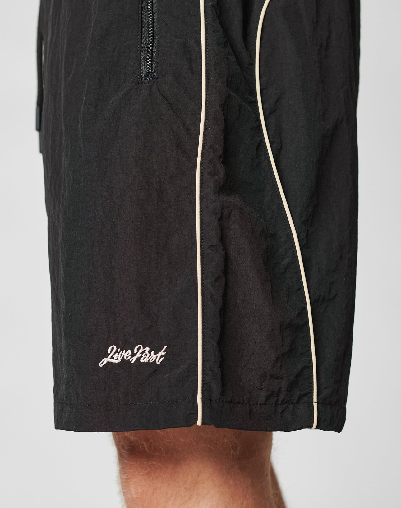 A close-up of a person wearing LIVE FAST DIE YOUNG Beach Shorts in black with white piping, featuring a mesh inlay and “Live Fast” embroidered in white near the hem.