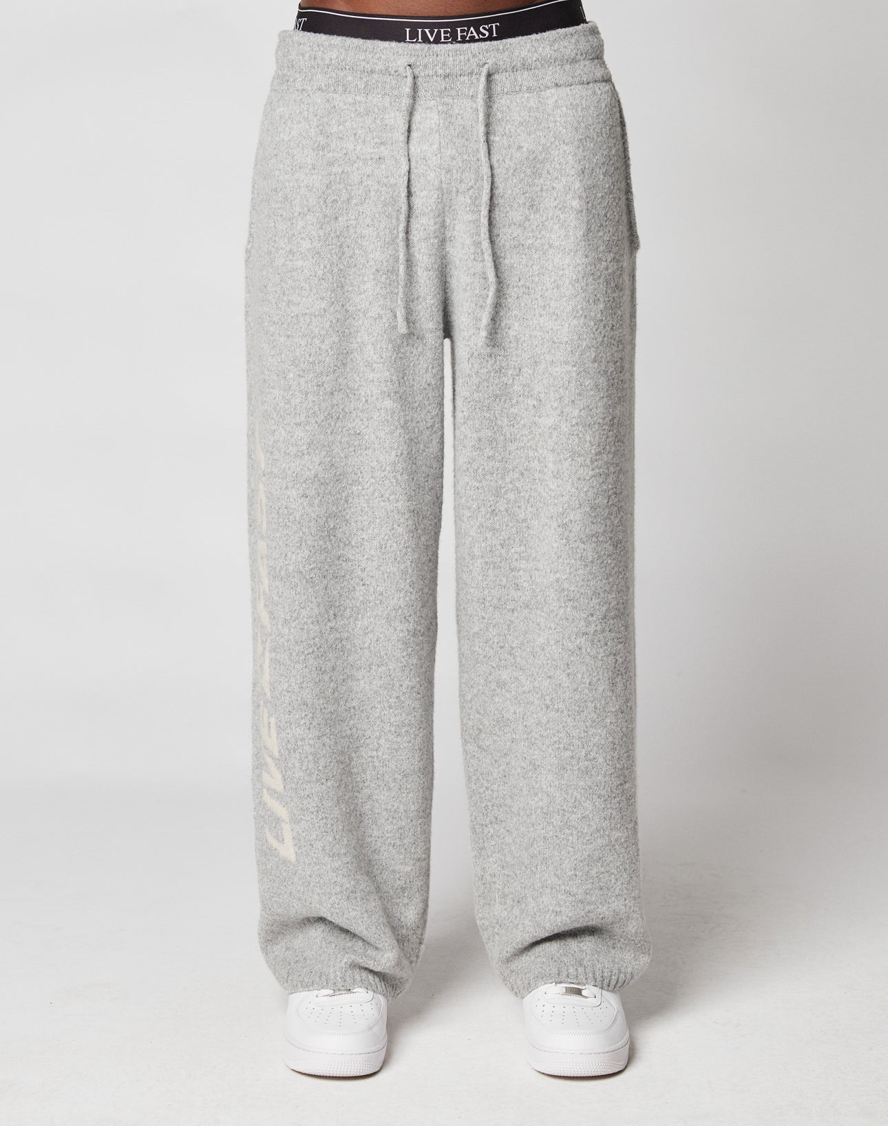 Wearing LIVE FAST DIE YOUNGs Live Fast Boucle Pants in light gray with an elastic waistband, wide fit, drawstrings, and white LIVE FAST accents, paired with white sneakers. Background is plain white.