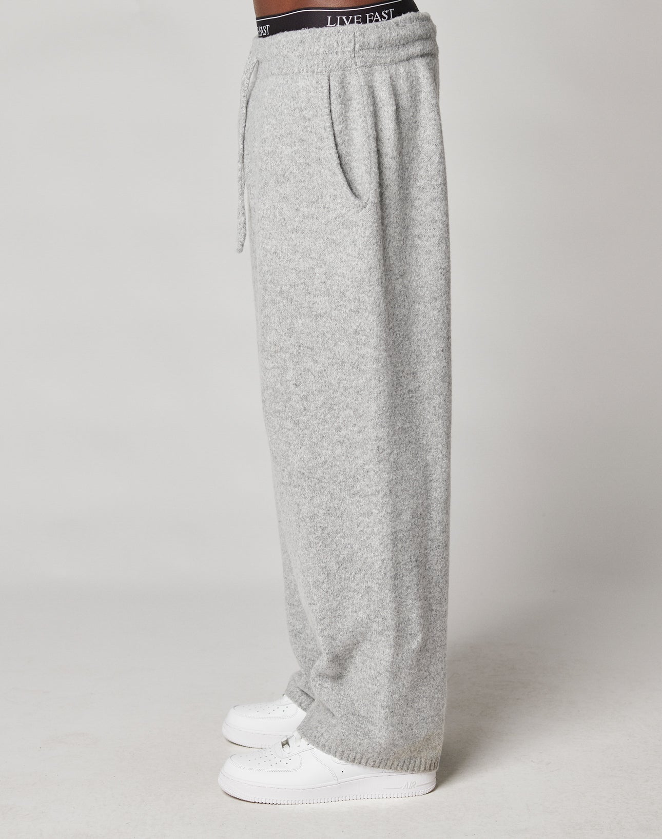 A person in profile wears LIVE FAST DIE YOUNG Live Fast Boucle Pants, wide fit and light gray with an elastic waistband and drawstring, paired with visible black LIVE FAST underwear and white sneakers, against a plain background.