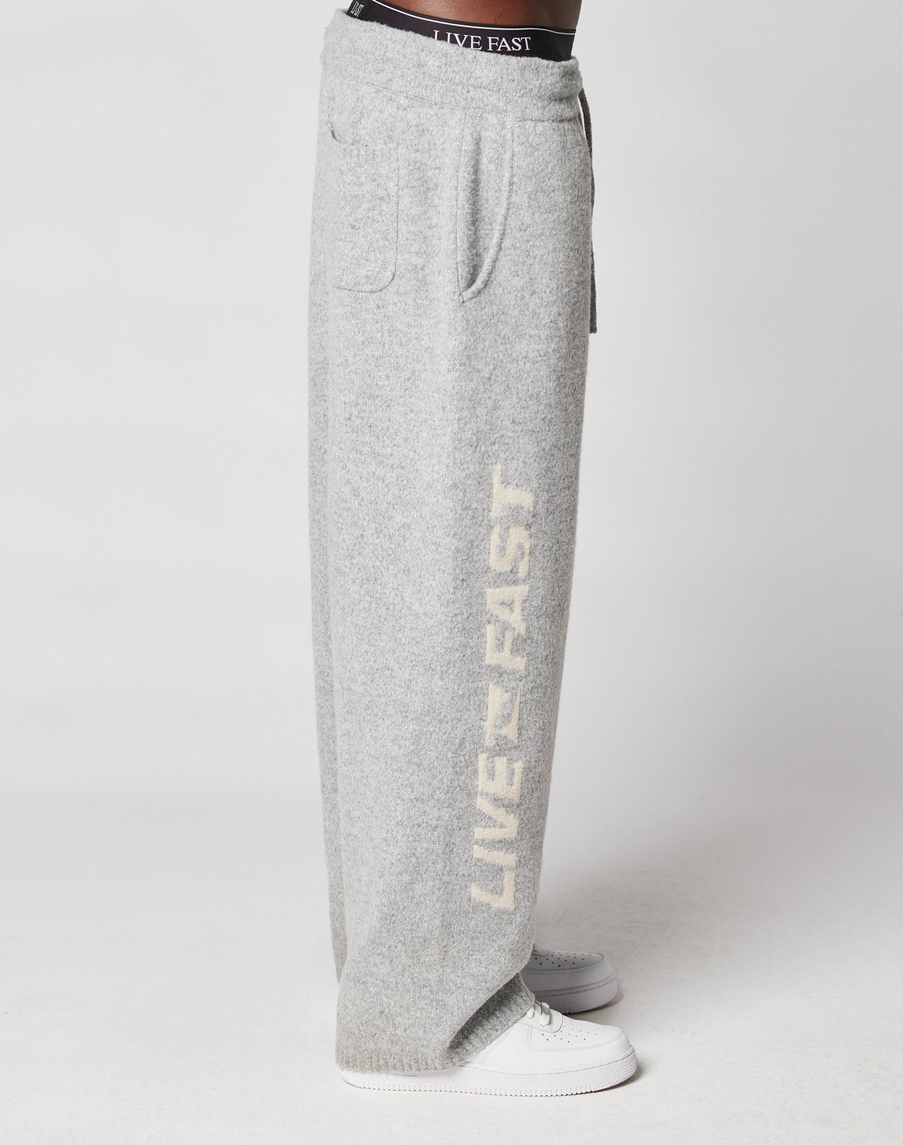 A person wears LIVE FAST DIE YOUNG’s Live Fast Boucle Pants—loose, light gray sweatpants with a comfy Elastik-Bund and white LIVE FAST text down the side—paired with white sneakers and black underwear visible above the wide waistband.
