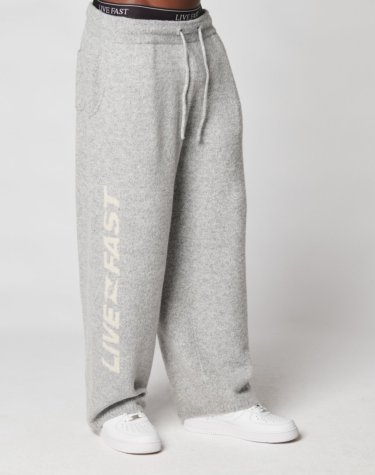 Someone wears loose-fitting, light gray Live Fast Boucle Pants by LIVE FAST DIE YOUNG, featuring an elastic waistband and LIVE FAST in white on one leg, styled with white sneakers and a visible black LIVE FAST waistband.