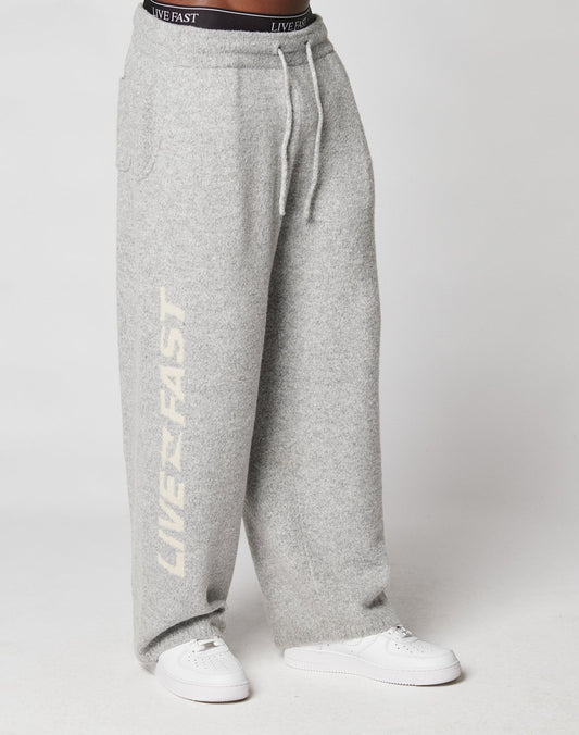 Someone wears loose-fitting, light gray Live Fast Boucle Pants by LIVE FAST DIE YOUNG, featuring an elastic waistband and LIVE FAST in white on one leg, styled with white sneakers and a visible black LIVE FAST waistband.