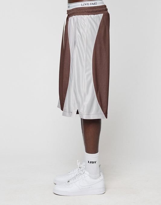 A person stands sideways in brown and white LFDY League Shorts by LIVE FAST DIE YOUNG, with white LEDY socks and white Nike sneakers. Only their lower half is visible against a plain background.