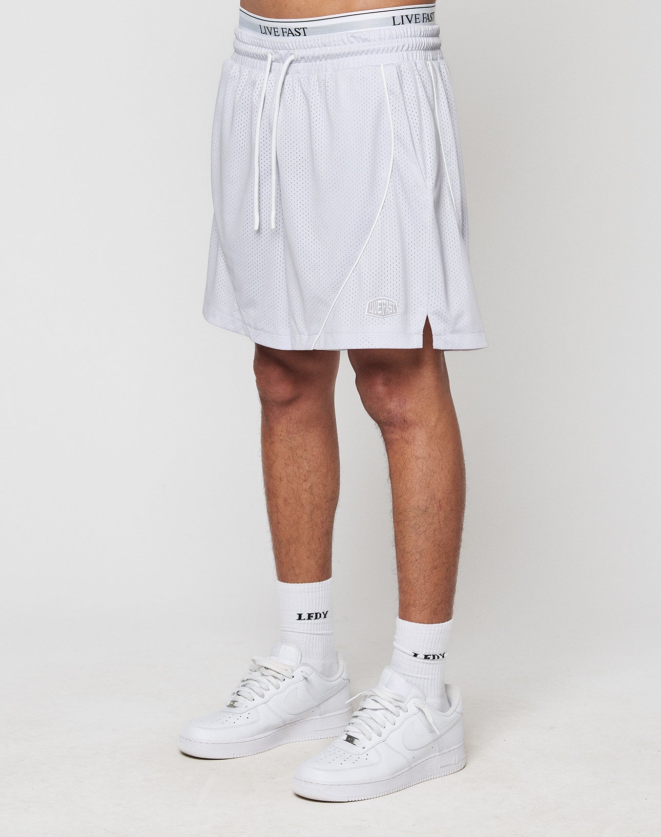 A person in white LIVE FAST DIE YOUNG LFDY Piping Mesh Shorts with a regular fit, elastic waistband, drawstring, and small logo near the hem, paired with white LFDTY socks and sneakers, stands against a plain light background. | model