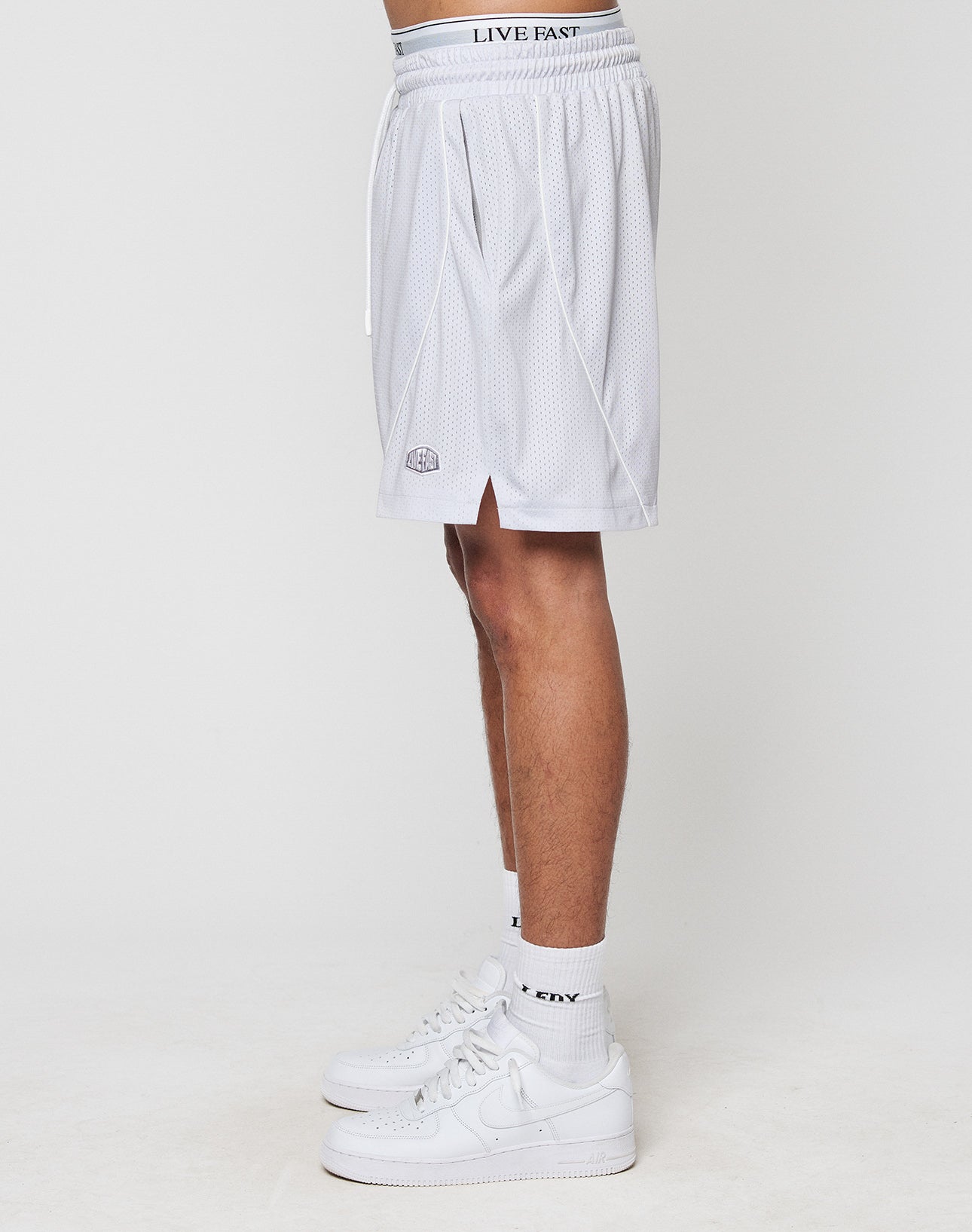 A person stands sideways in LFDY Piping Mesh Shorts by LIVE FAST DIE YOUNG—regular fit, white polyester with small side slit and elastic drawstring waist—paired with white crew socks and sneakers, against a plain white background.