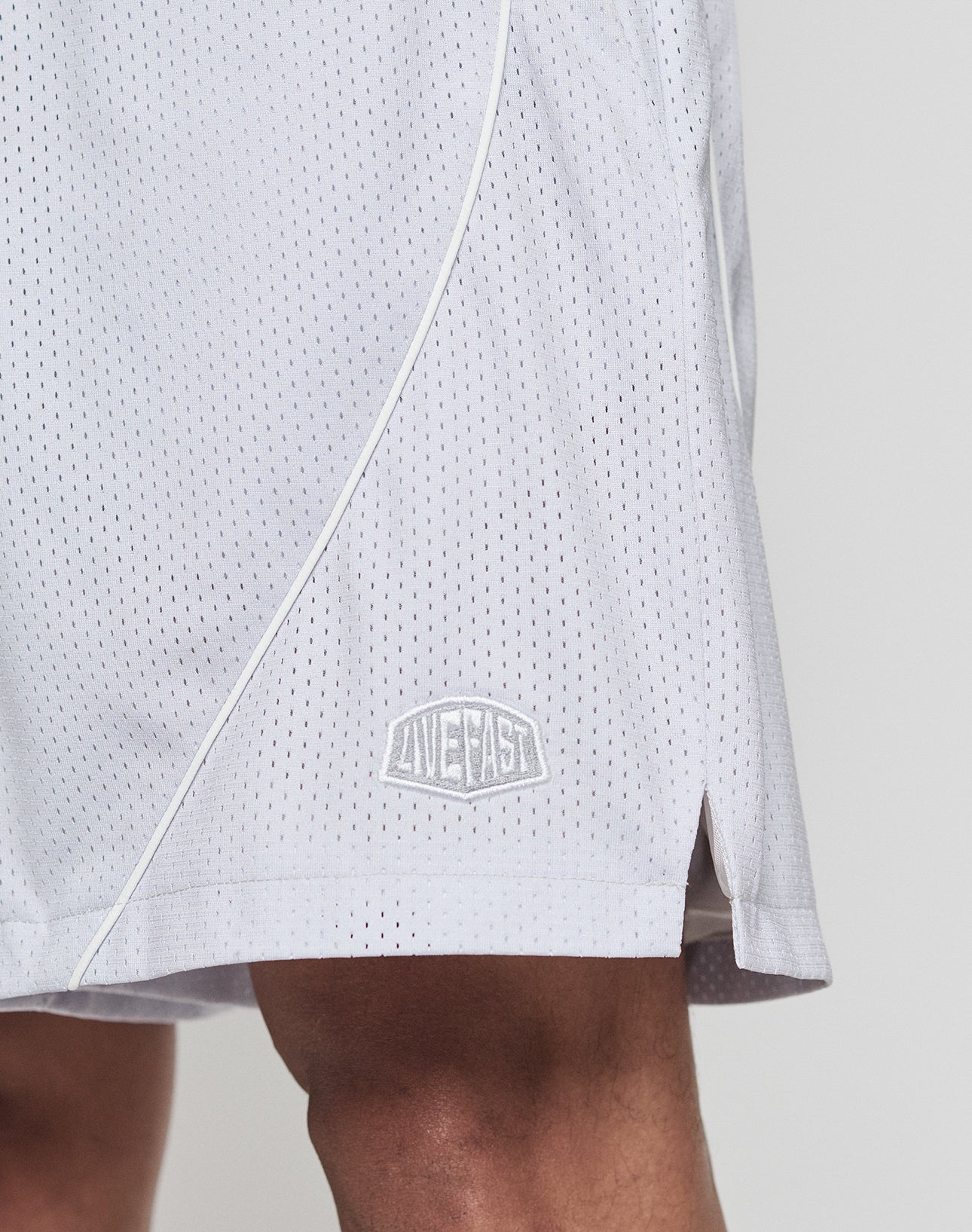 A close-up of a person wearing LFDY Piping Mesh Shorts by LIVE FAST DIE YOUNG, featuring white mesh fabric, a small embroidered logo near the hem, a side slit, and an elastic waistband for comfort.