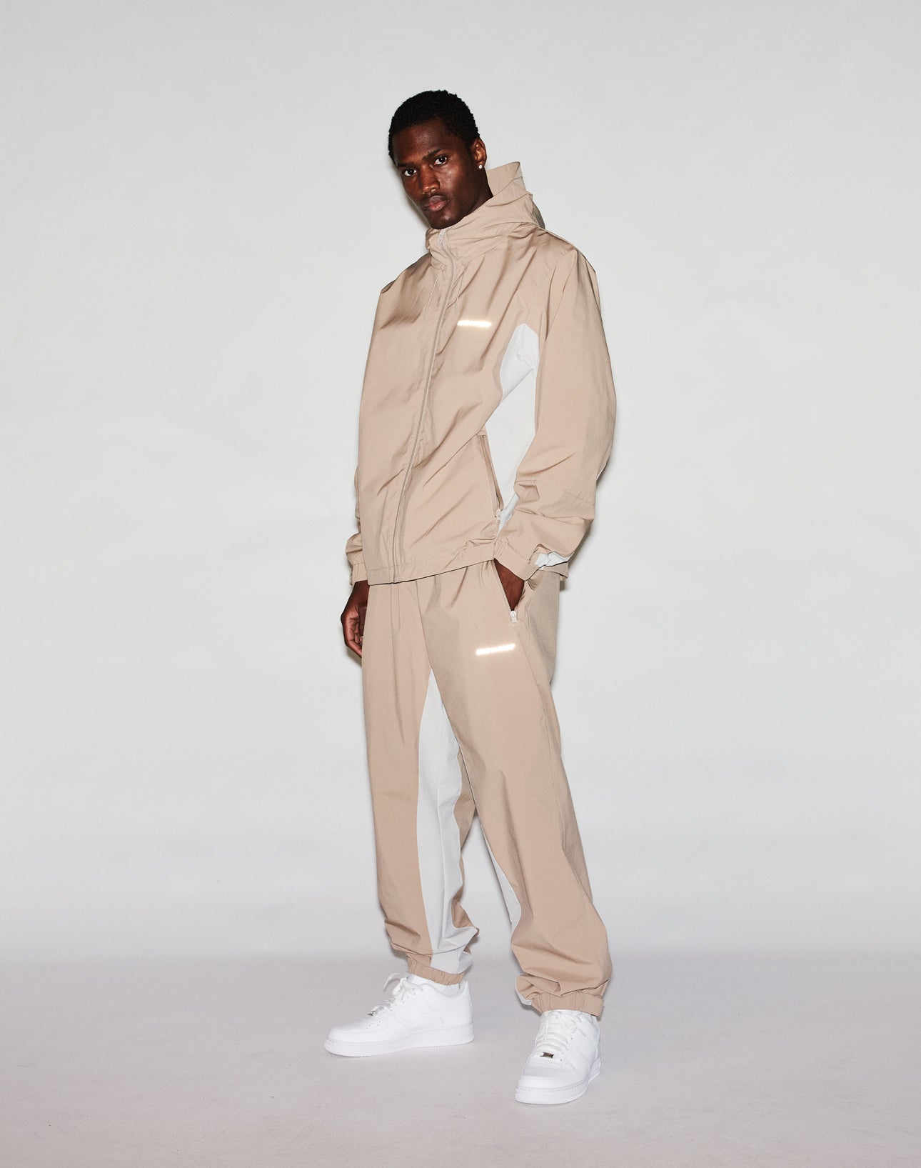A man stands against a plain background wearing LIVE FAST DIE YOUNG Basic Shell Pants with an elastic waistband and drawstring, paired with a beige and white oversize windbreaker jacket and white sneakers. He faces the camera with a neutral expression.