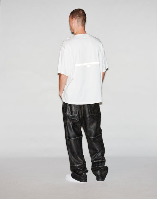 A person faces away from the camera, wearing a white LFDY Trinity Tee by LIVE FAST DIE YOUNG and loose black leather-like pants, against a plain light background.