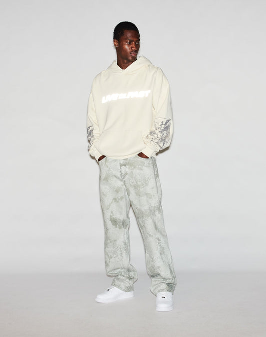 A man wears the LFDY Window Hooded by LIVE FAST DIE YOUNG, featuring “LATE FAST” reflective print, logo embroidery, patterned sleeves, light grey patterned pants, and white sneakers. He looks down left for an oversized fit style.