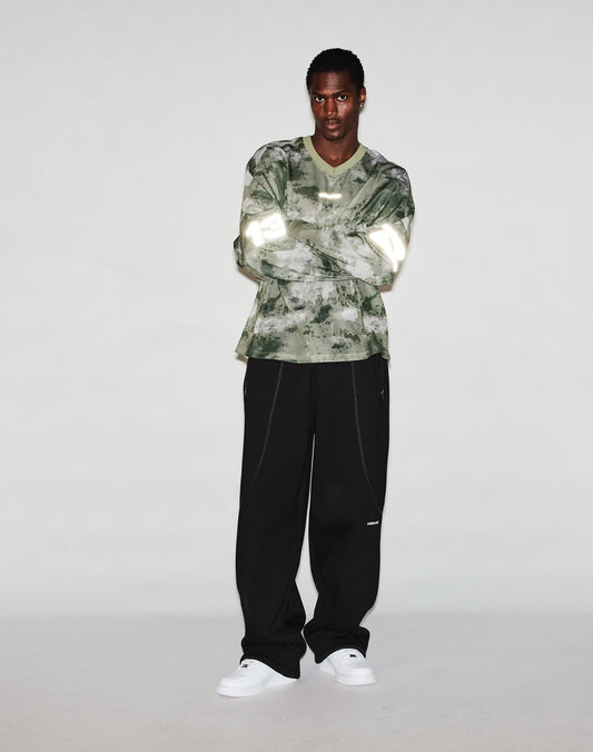 A person stands against a plain light background, wearing the LIVE FAST DIE YOUNG Camo Mesh Jersey in an oversize fit, loose black pants, and white sneakers. Their arms are crossed as they face the camera.