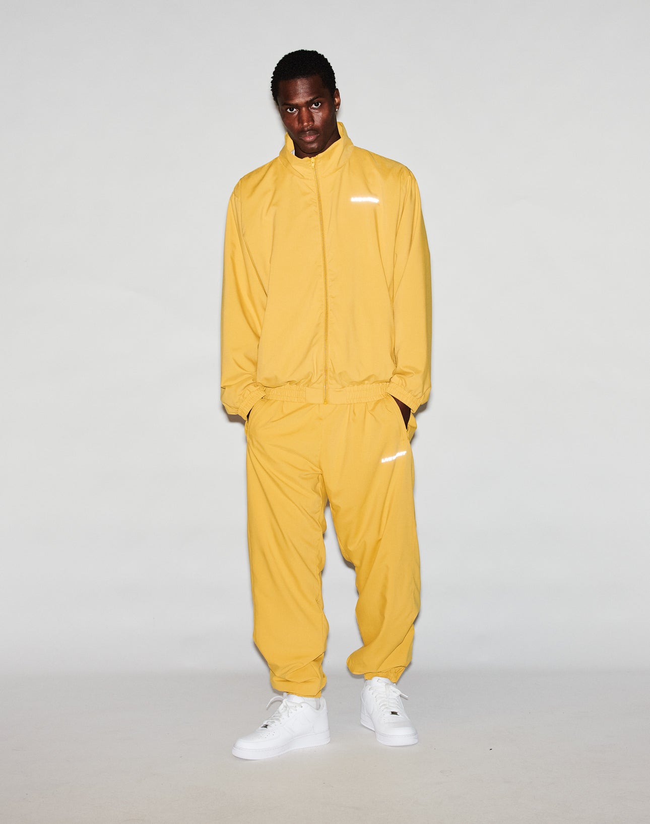 A person stands against a plain background, wearing the yellow LFDY Trackjacket by LIVE FAST DIE YOUNG with an oversize fit, reflective print, and white sneakers, looking directly at the camera with hands in pockets.