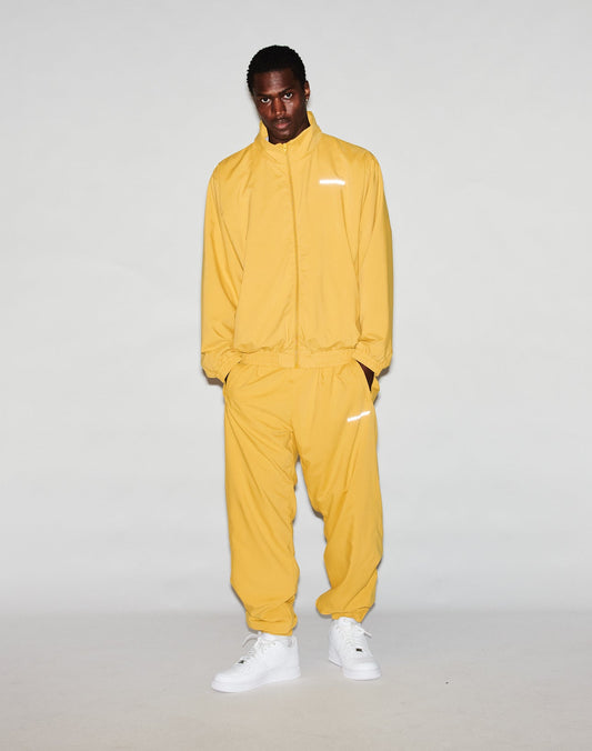 A person stands against a plain background, wearing the yellow LFDY Trackjacket by LIVE FAST DIE YOUNG with an oversize fit, reflective print, and white sneakers, looking directly at the camera with hands in pockets.