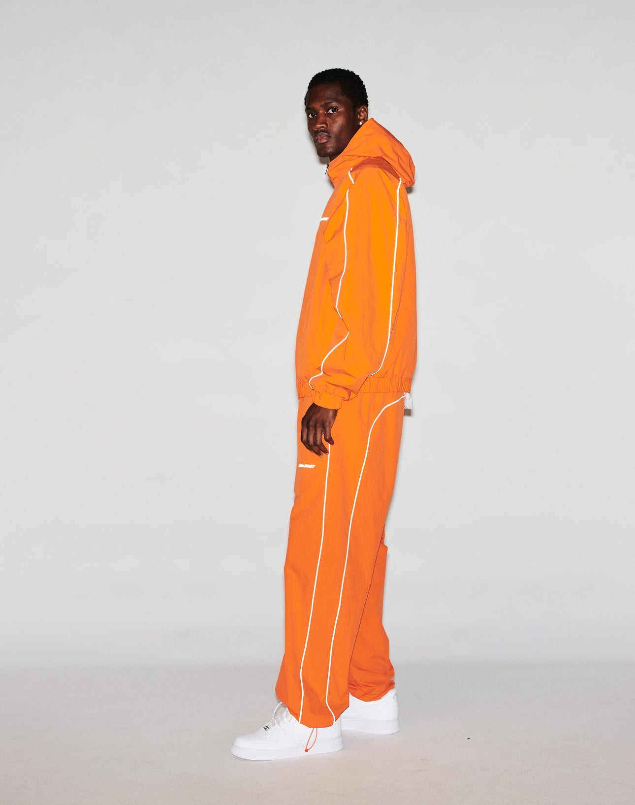 A person stands sideways against a plain background, wearing the orange Live Fast Trackpants by LIVE FAST DIE YOUNG with white piping and Breiter Elastik-Bund. The hood is up, they face the camera. White sneakers and a reflektierender Logo Print finish the look.