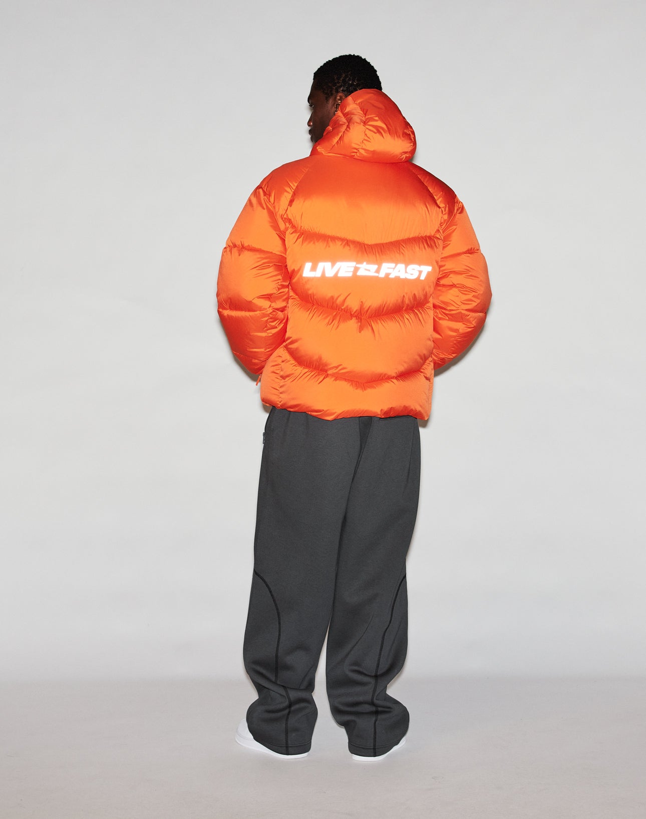A person faces away in the LIVE FAST DIE YOUNG Live Fast Puffer Jacket—bright orange with a reflective LIVE FAST print on the back—styled with loose dark gray pants and white shoes, emphasizing the jacket’s oversize fit against a plain background.