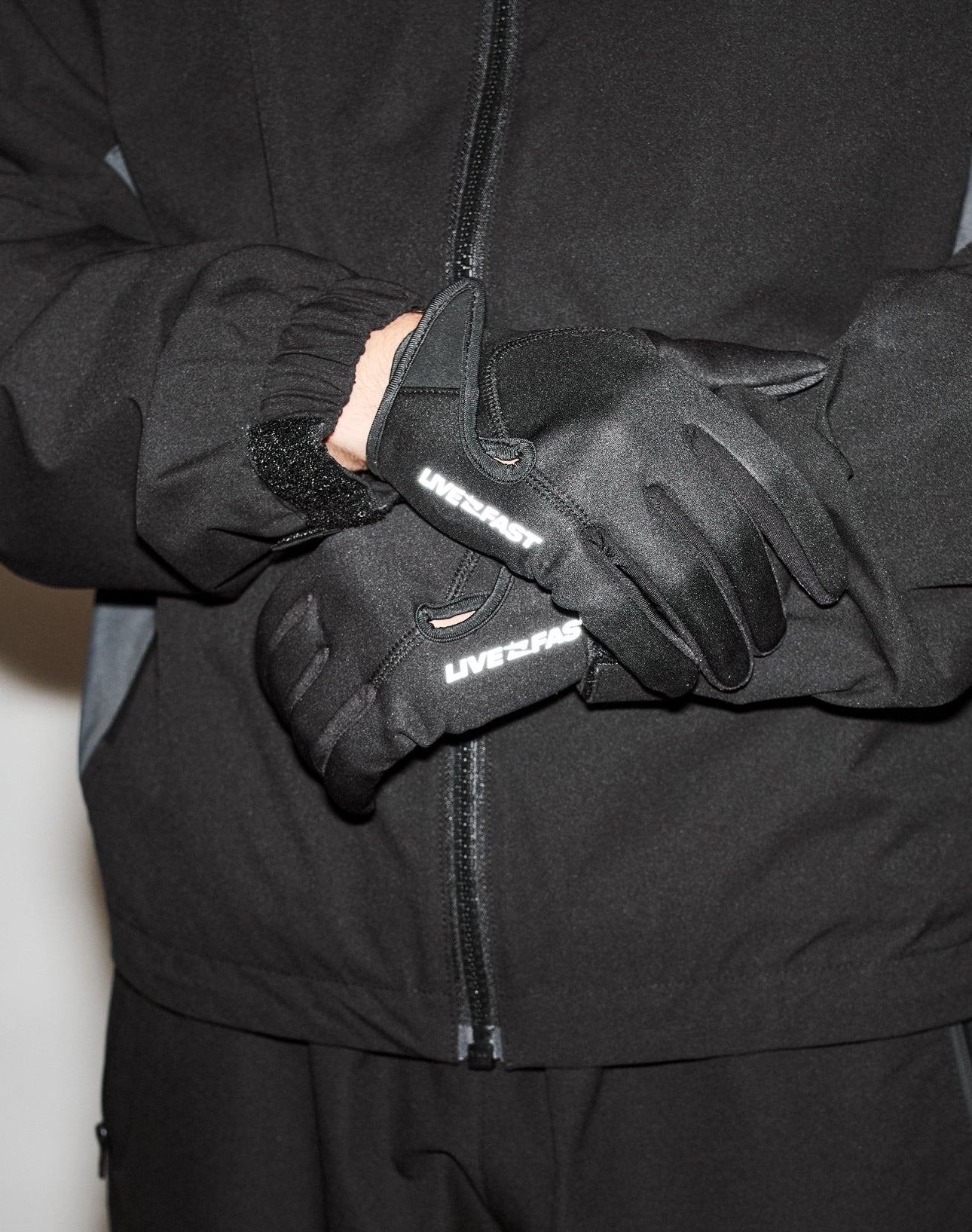 Someone wearing LIVE FAST DIE YOUNG’s Live Fast Gloves, which have reflective print, crosses their gloved hands in front of a black zip-up jacket and pants.