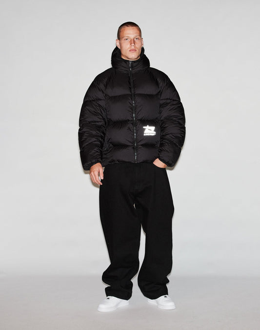 A person stands against a plain background wearing the LIVE FAST DIE YOUNG Live Fast Puffer Jacket in black, loose black pants, and white sneakers. The outfit is casual and warm, and the individual faces forward with arms relaxed at their sides.