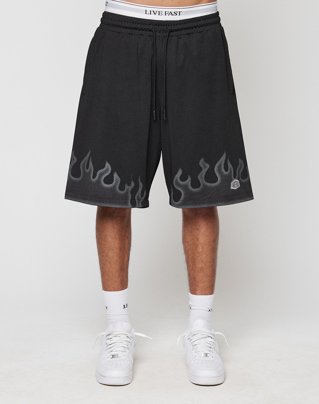 A person wears LIVE FAST DIE YOUNG Burning Mesh Shorts with gray flame print along the hem, LIVE FAST on the elastic waistband, paired with white socks and sneakers. Photo shows a plain background.