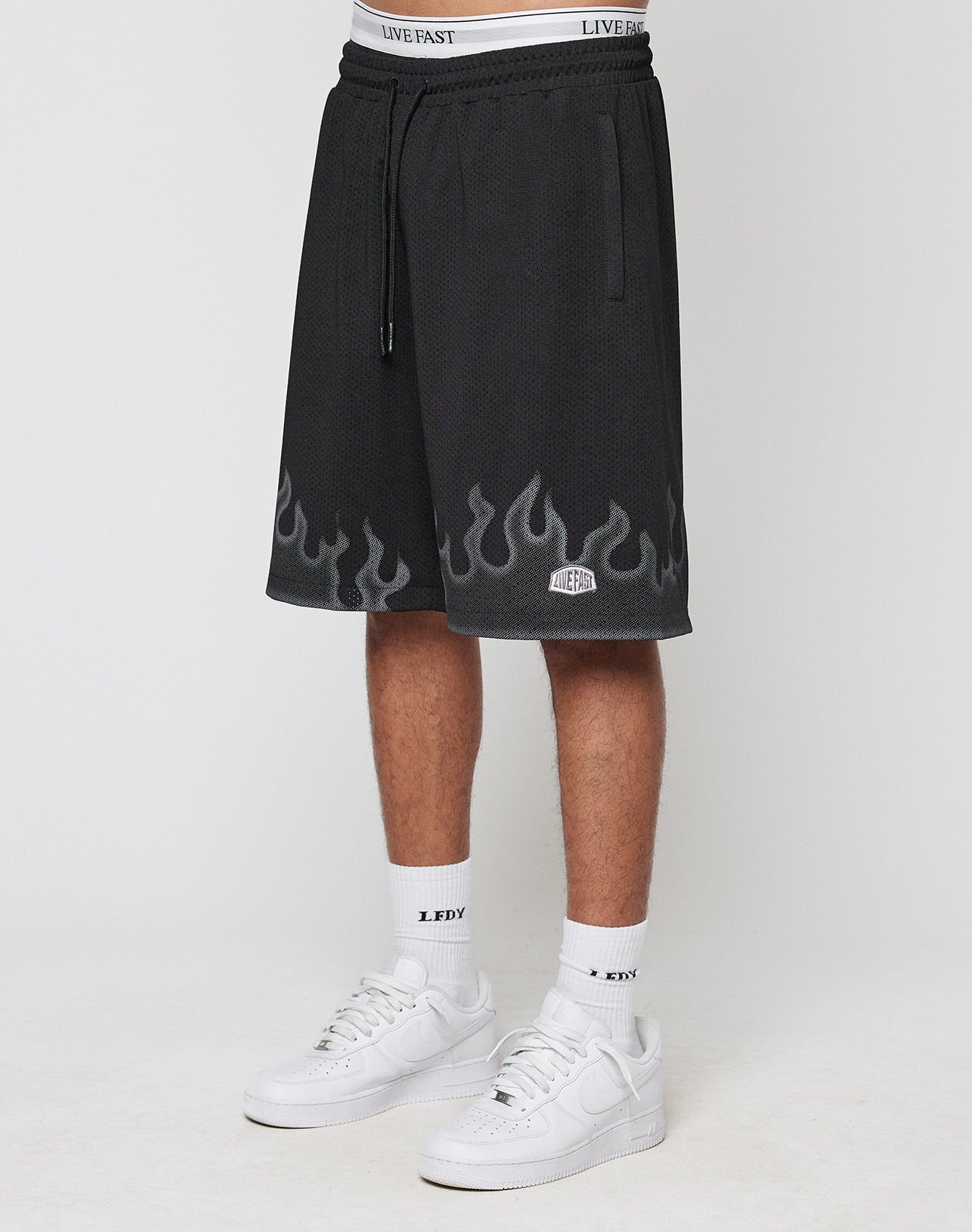 A person wears LIVE FAST DIE YOUNG Burning Mesh Shorts with a Flammenprint hem and elastic waistband, showing the white underwears band above. Paired with white socks and sneakers, theyre photographed against a plain background. | model