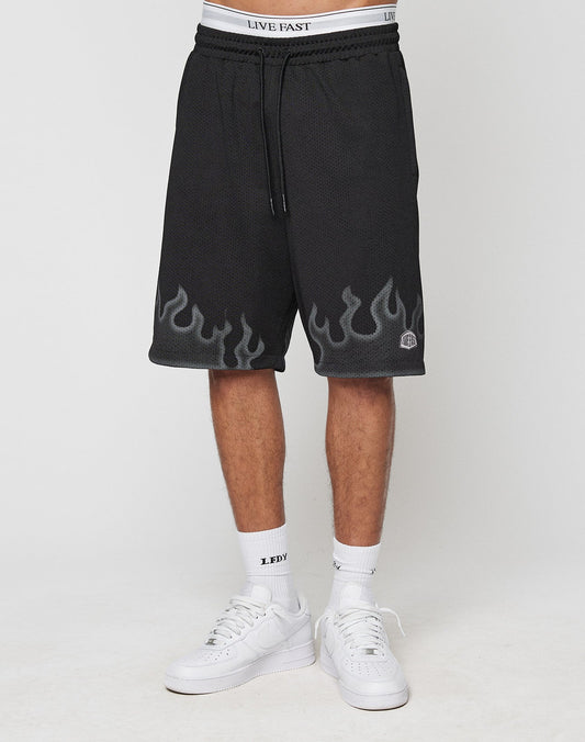 A person wears LIVE FAST DIE YOUNG Burning Mesh Shorts with a gray Flammenprint and LIVE FAST waistband, white LEDY socks, and white sneakers, standing against a plain white background.
