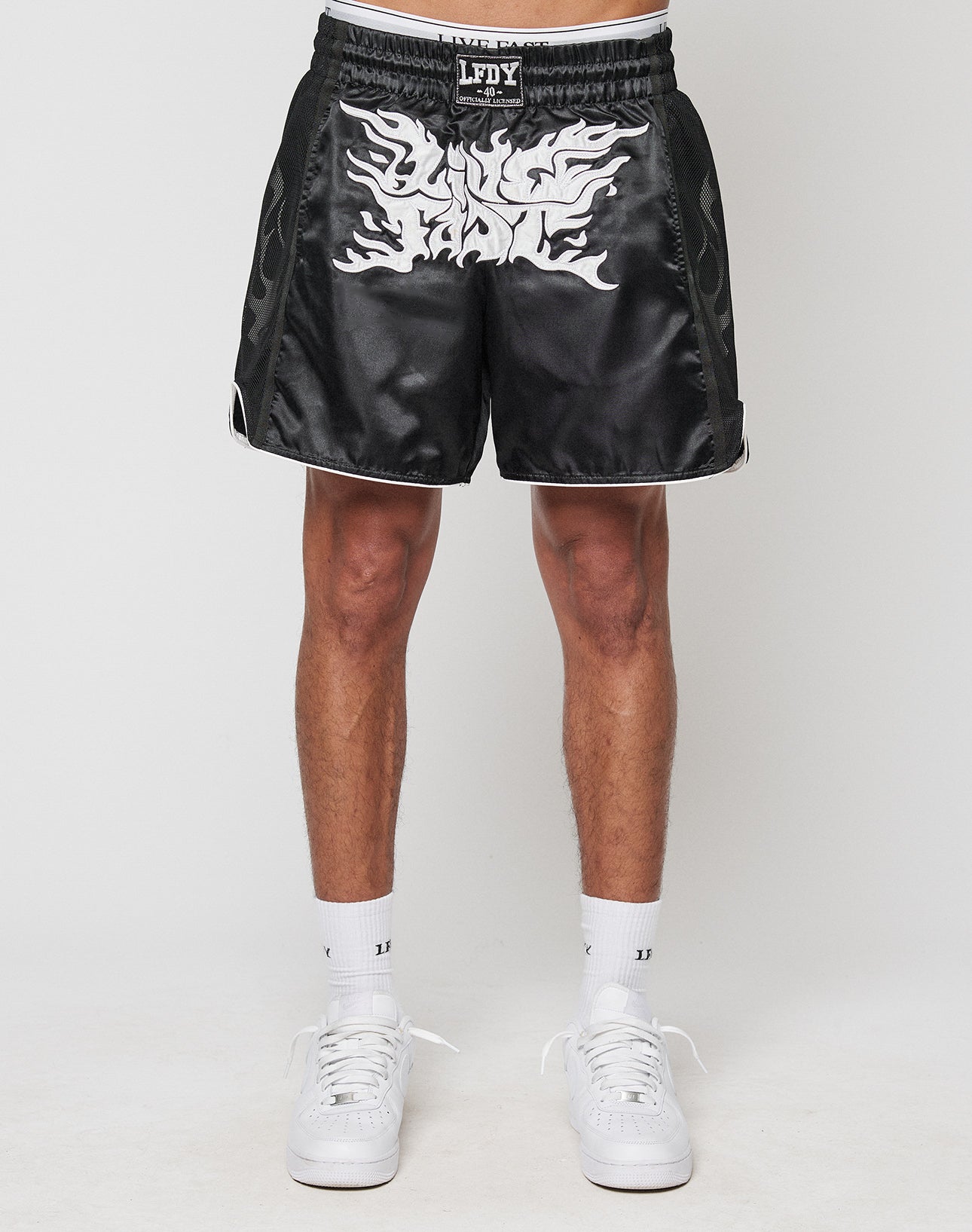 A person wears LIVE FAST DIE YOUNGs LFDY Fighting Shorts—regular fit black satin with white flame graphics and elastic waistband—paired with white socks and sneakers, standing against a plain white background.