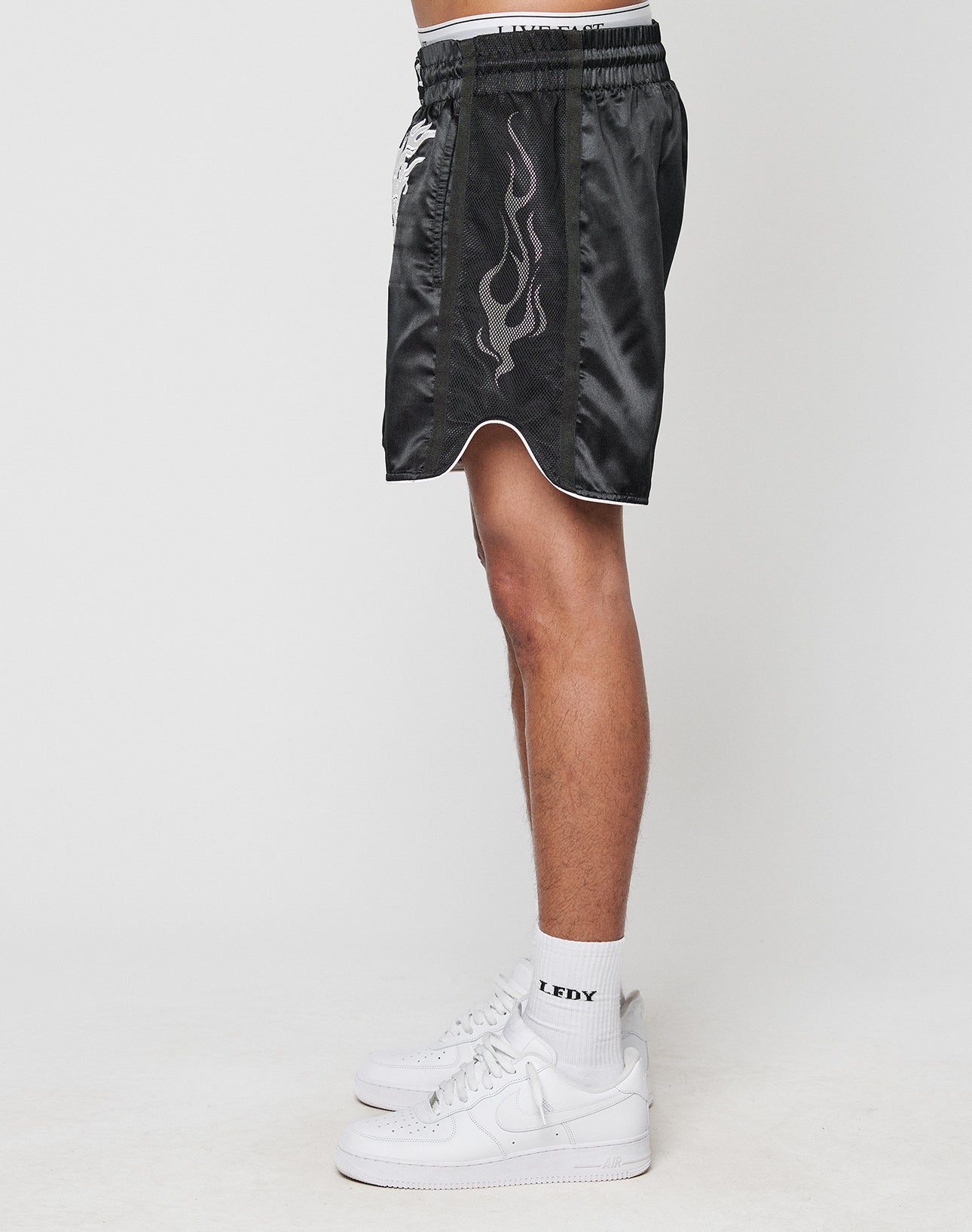 A person stands sideways wearing LIVE FAST DIE YOUNG’s LFDY Fighting Shorts—black regular fit with flame design and elastic waistband—plus white LFDY crew socks and white sneakers, all against a plain white background.