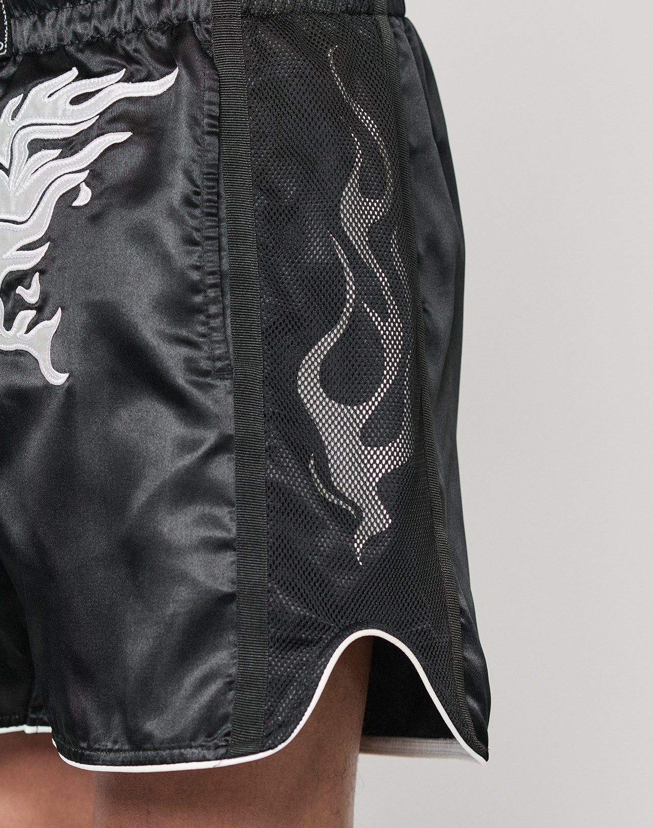 Close-up of the left side of a person wearing LIVE FAST DIE YOUNG LFDY Fighting Shorts in black with a white flame design, mesh side panel, and elastic waistband, revealing part of the leg and hip against a plain background.