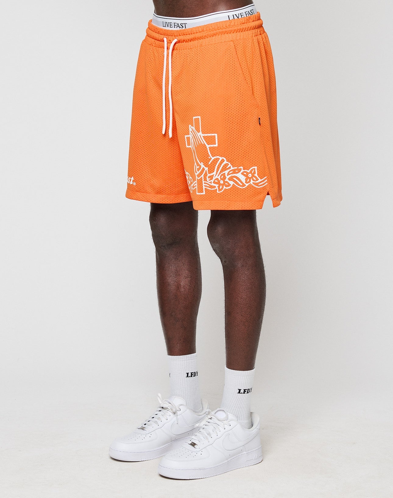 A person wearing LIVE FAST DIE YOUNG LFDY Praying Mesh Shorts in orange with white graphics, white socks, and Nike Air Force 1 sneakers stands against a plain white background. Only the lower half of the body is visible. | model