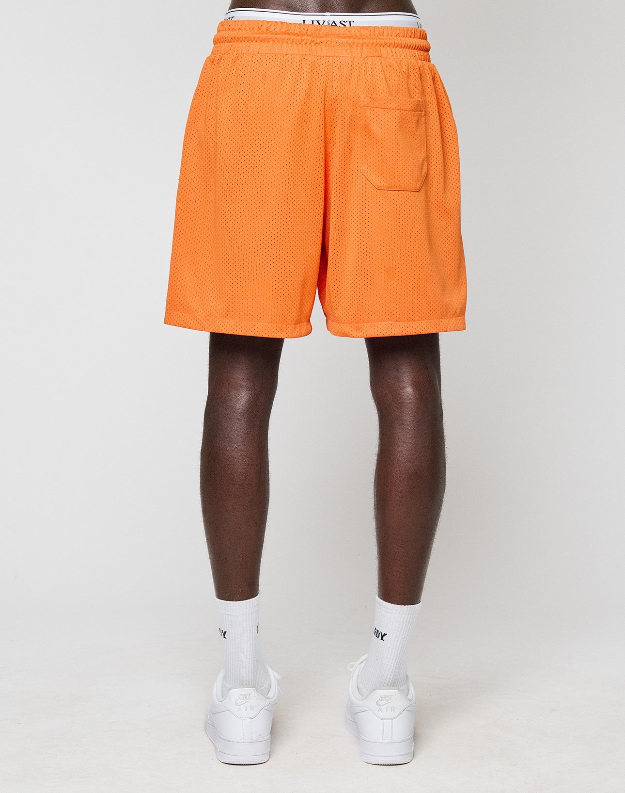 A person stands with their back to the camera in orange LFDY Praying Mesh Shorts by LIVE FAST DIE YOUNG, featuring a Regular Fit, elastic waistband, small back pocket, and visible underwear band. They wear white socks and sneakers against a plain white background.