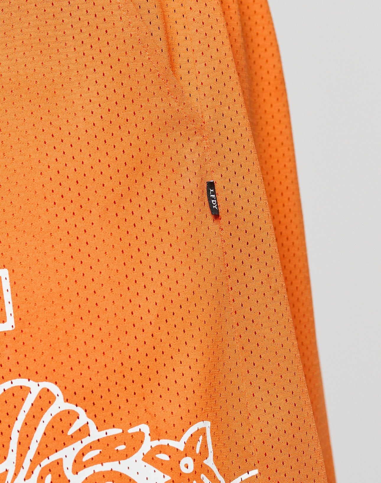 Close-up of orange LFDY Praying Mesh Shorts by LIVE FAST DIE YOUNG, showing perforated polyester fabric with an elastic waistband, small black LFDY tag, and part of a white graphic near the hem.