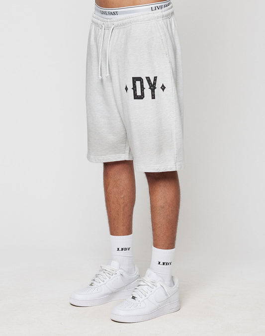 A person in LIVE FAST DIE YOUNG DY Rivet Shorts (light gray, 100% Baumwolle) with DY on the left leg, white LFDY socks, and white Nike sneakers stands against a plain white background. | model