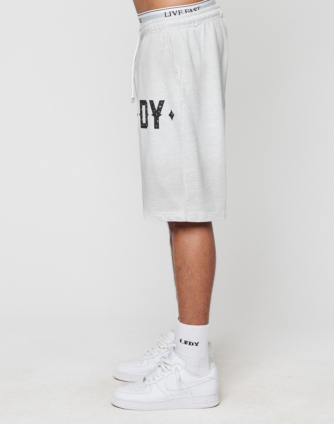 A person stands sideways wearing light gray, oversize fit DY Rivet Shorts by LIVE FAST DIE YOUNG, featuring EDY text in black, matching socks with EDY, white Nike sneakers, and a waistband reading LIVE FAST. Plain white background.