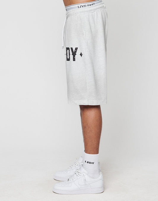 A person stands sideways wearing light gray, oversize fit DY Rivet Shorts by LIVE FAST DIE YOUNG, featuring EDY text in black, matching socks with EDY, white Nike sneakers, and a waistband reading LIVE FAST. Plain white background.