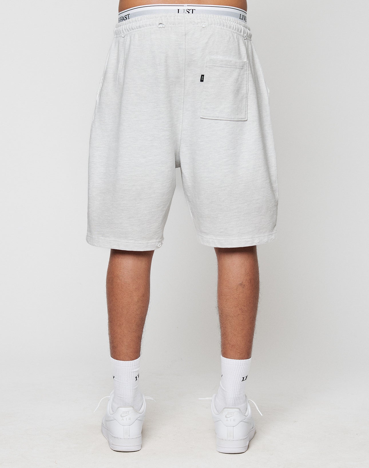 A person faces away, wearing LIVE FAST DIE YOUNGs DY Rivet Shorts in light gray with a back pocket and LEISURE-print waistband. Made from 100% Baumwolle, theyre paired with white socks and sneakers for comfort and style.