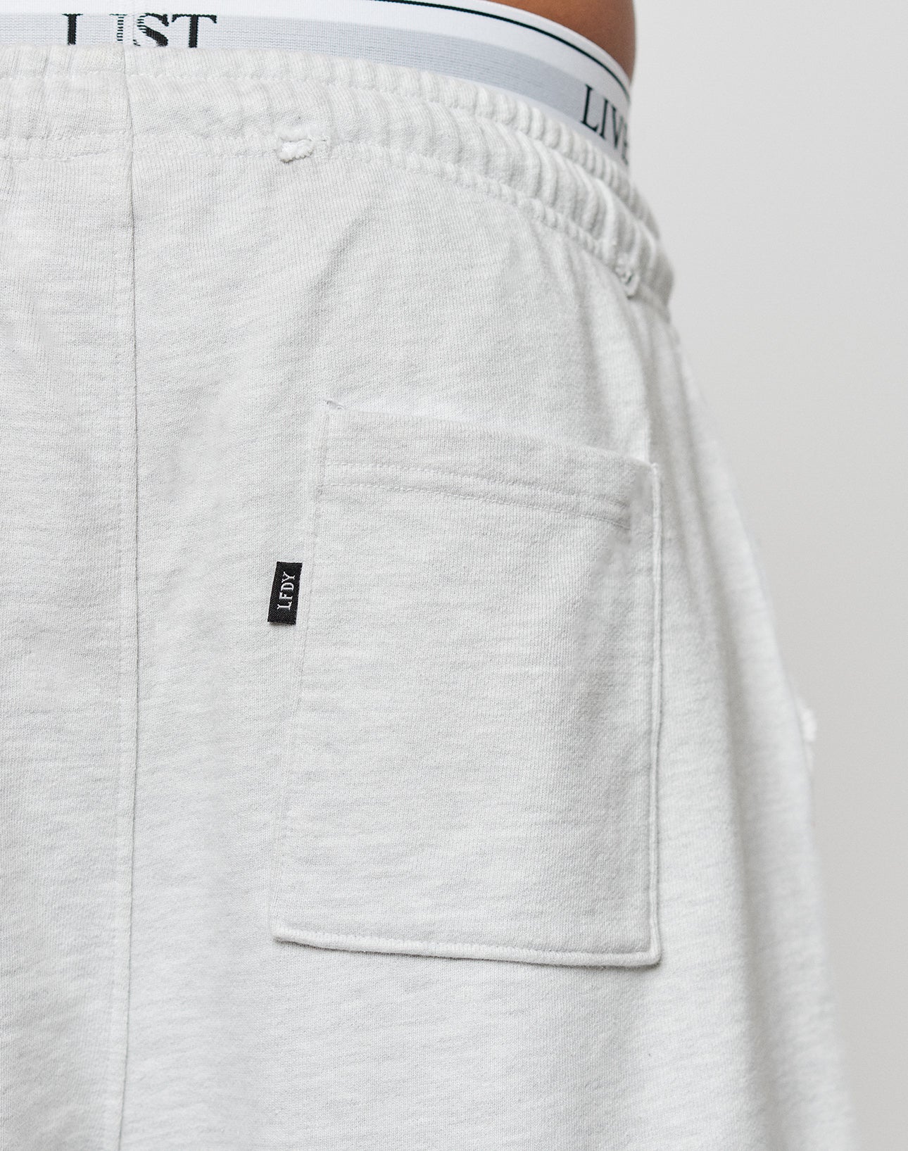 Close-up of the back pocket of light gray DY Rivet Shorts by LIVE FAST DIE YOUNG, featuring a black LIST tag on the pocket seam and white 100% Baumwolle underwear with LIST branding visible above the waistband.