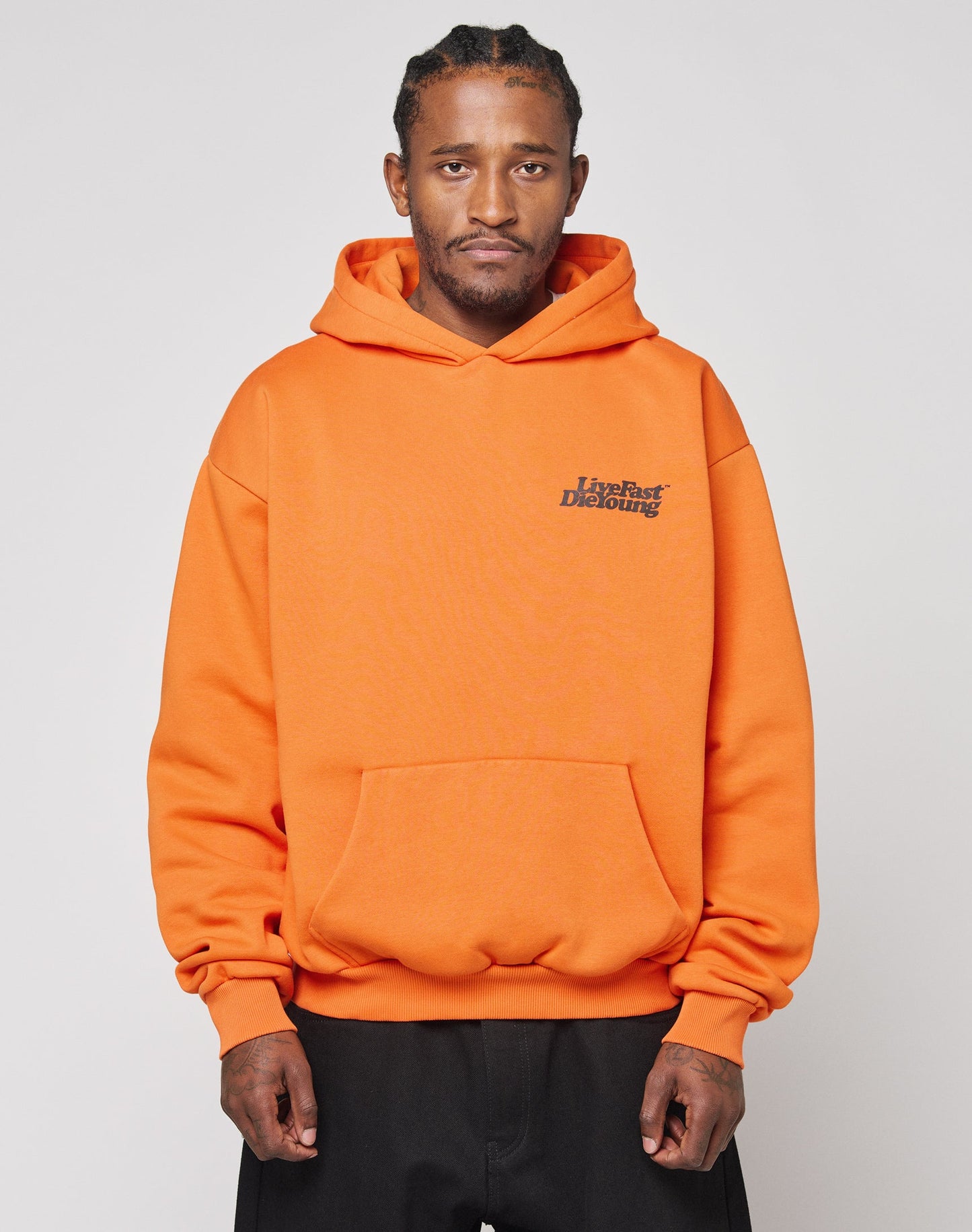 A man stands facing forward against a plain light background, wearing the LIVE FAST DIE YOUNG Basic Orange Hooded sweatshirt made from Baumwolle and Polyester, featuring a small printed chest logo. His hands are by his sides with a neutral expression.